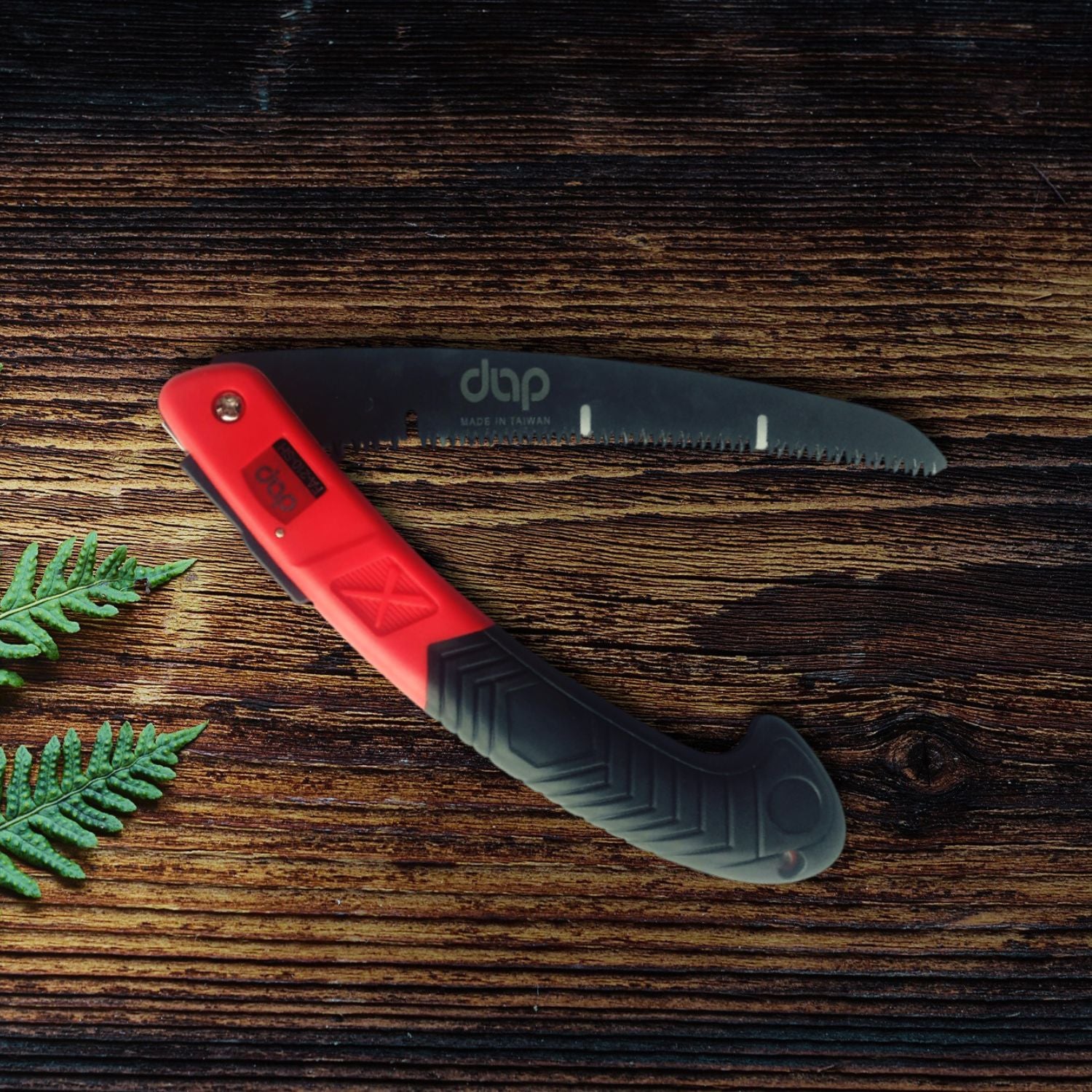 Folding saw with red handle and on a wooden surface with green leaves.
