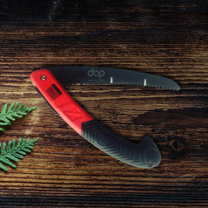 Folding saw with red handle and on a wooden surface with green leaves.