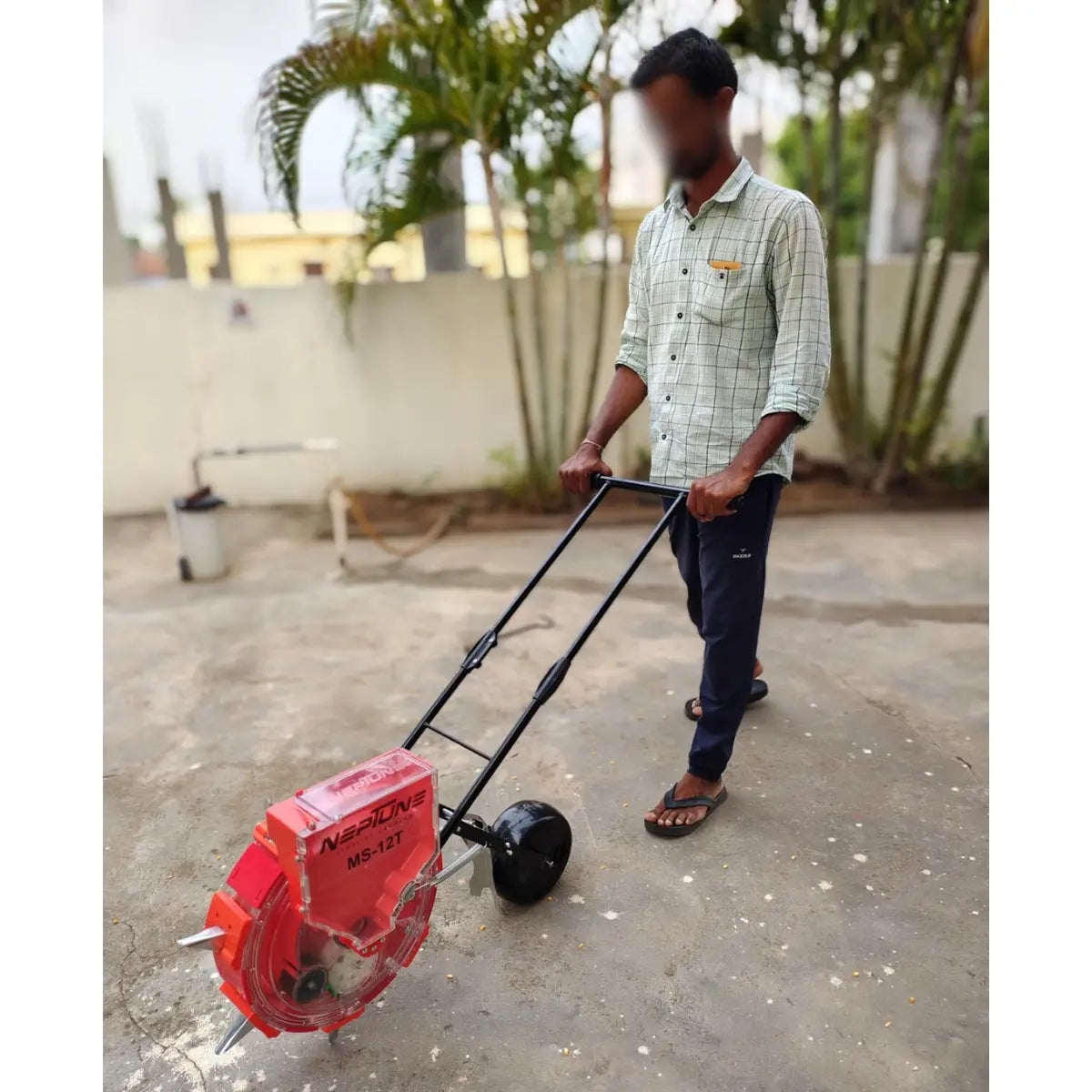 Person using a red manual seed sowing machine on a concrete surface with plants in the background